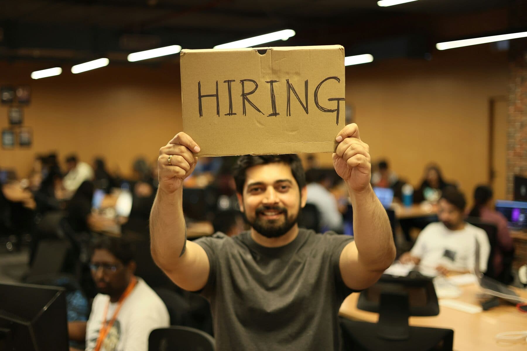 A recruiter smiling and holding a handwritten cardboard HIRING sign above their head in a room full of students working at laptops
